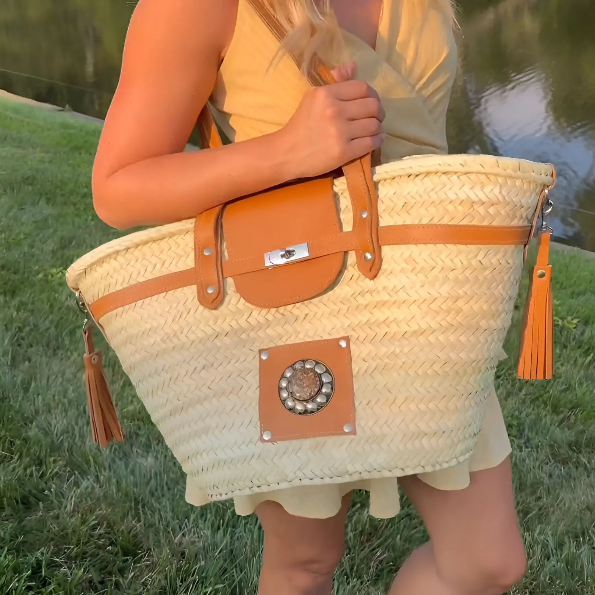 A person holding a large straw basket with leather straps over their shoulder, set against a grassy background.
