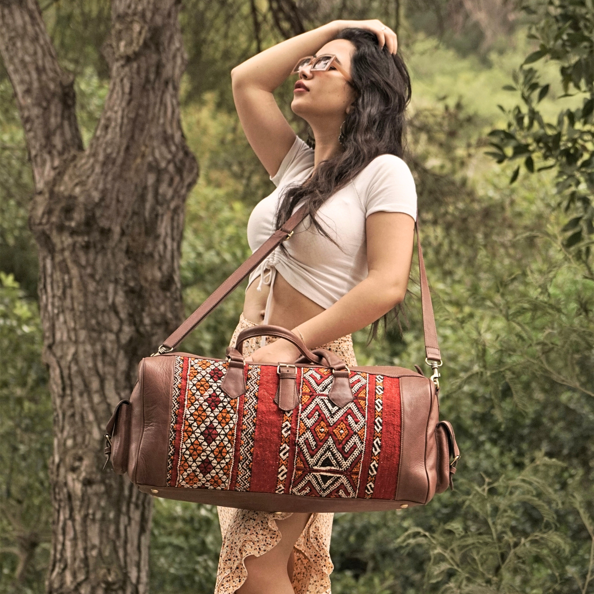 A person standing outdoors wearing a white top and carrying a brown leather duffel bag with a colorful kilim design.