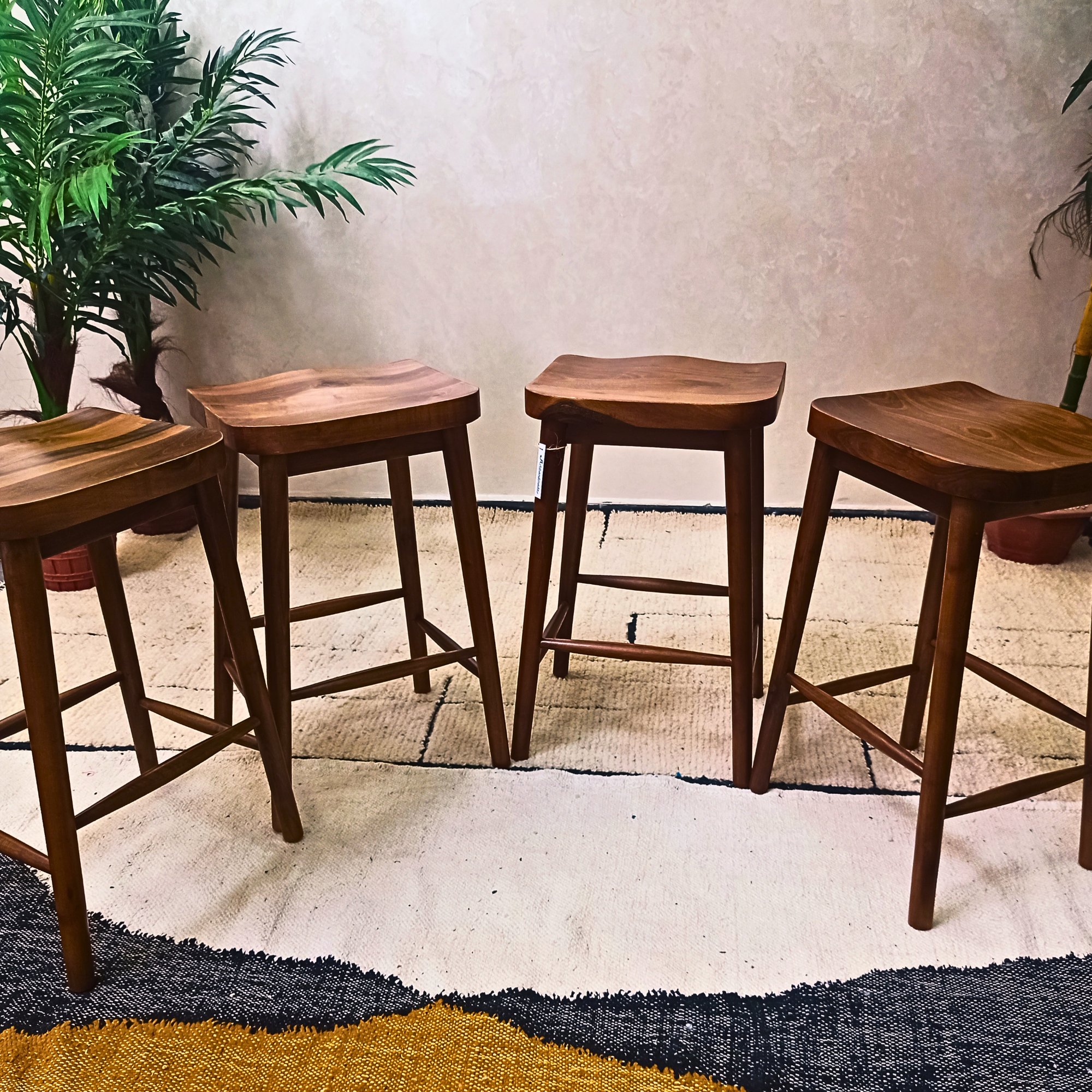 Four walnut wooden bar stools arranged on a textured floor with a plant in the background.