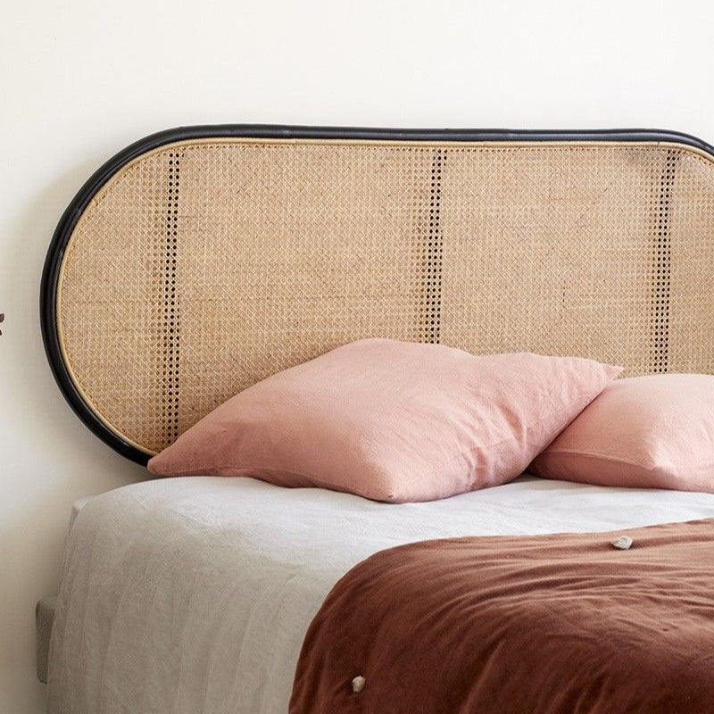A rattan headboard with a wood frame, mounted on a wall above a bed with pink pillows and a brown duvet cover. The room has a cozy ambiance with a side table, lamp, and decorative items.